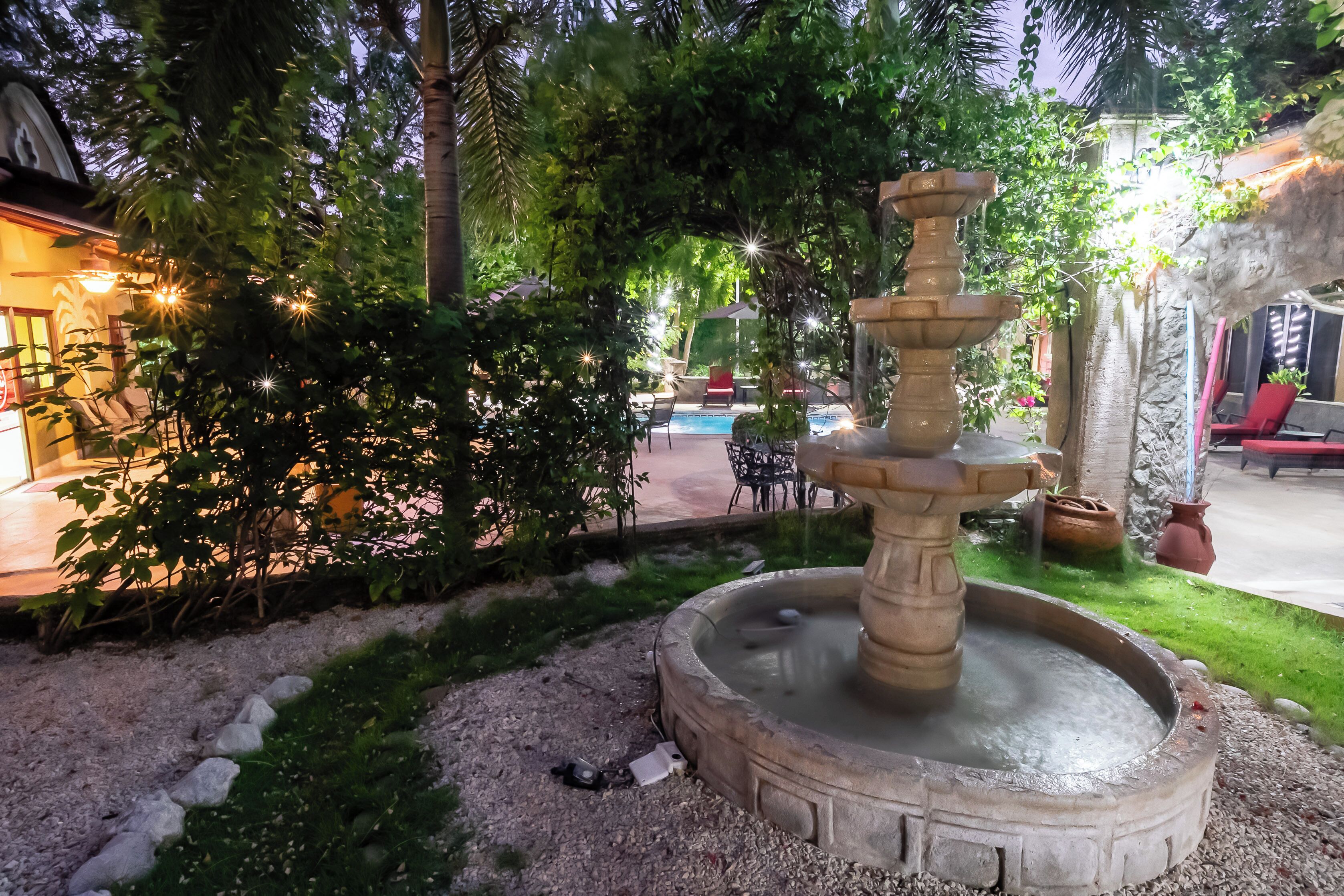 A fountain in a garden area with a pool and seating in the background.
