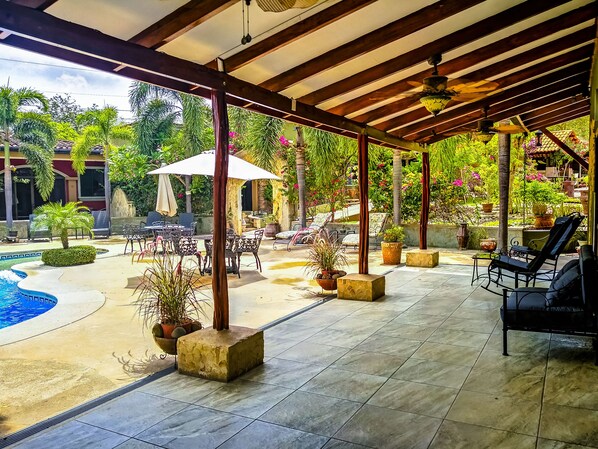 A covered outdoor area with tiled flooring, lounge chairs, and a pool.