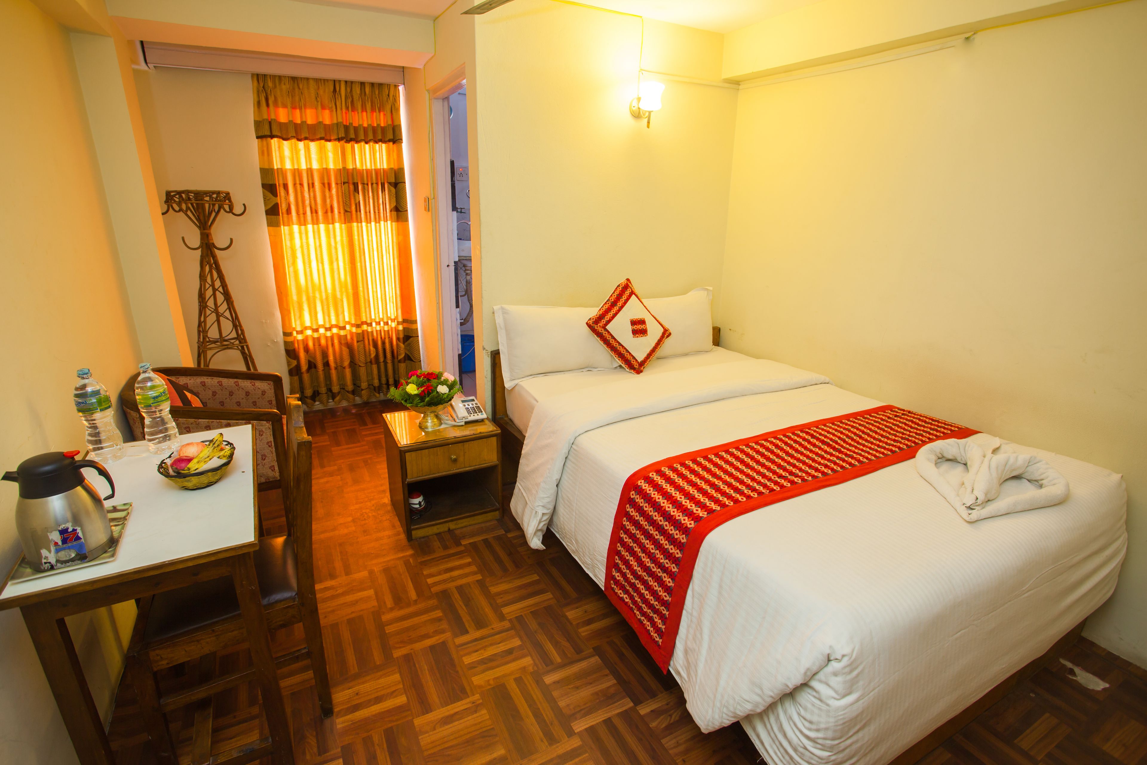 Una habitación de hotel con una cama, un escritorio con hervidor, una silla, una mesita de noche con teléfono y una ventana con cortinas.