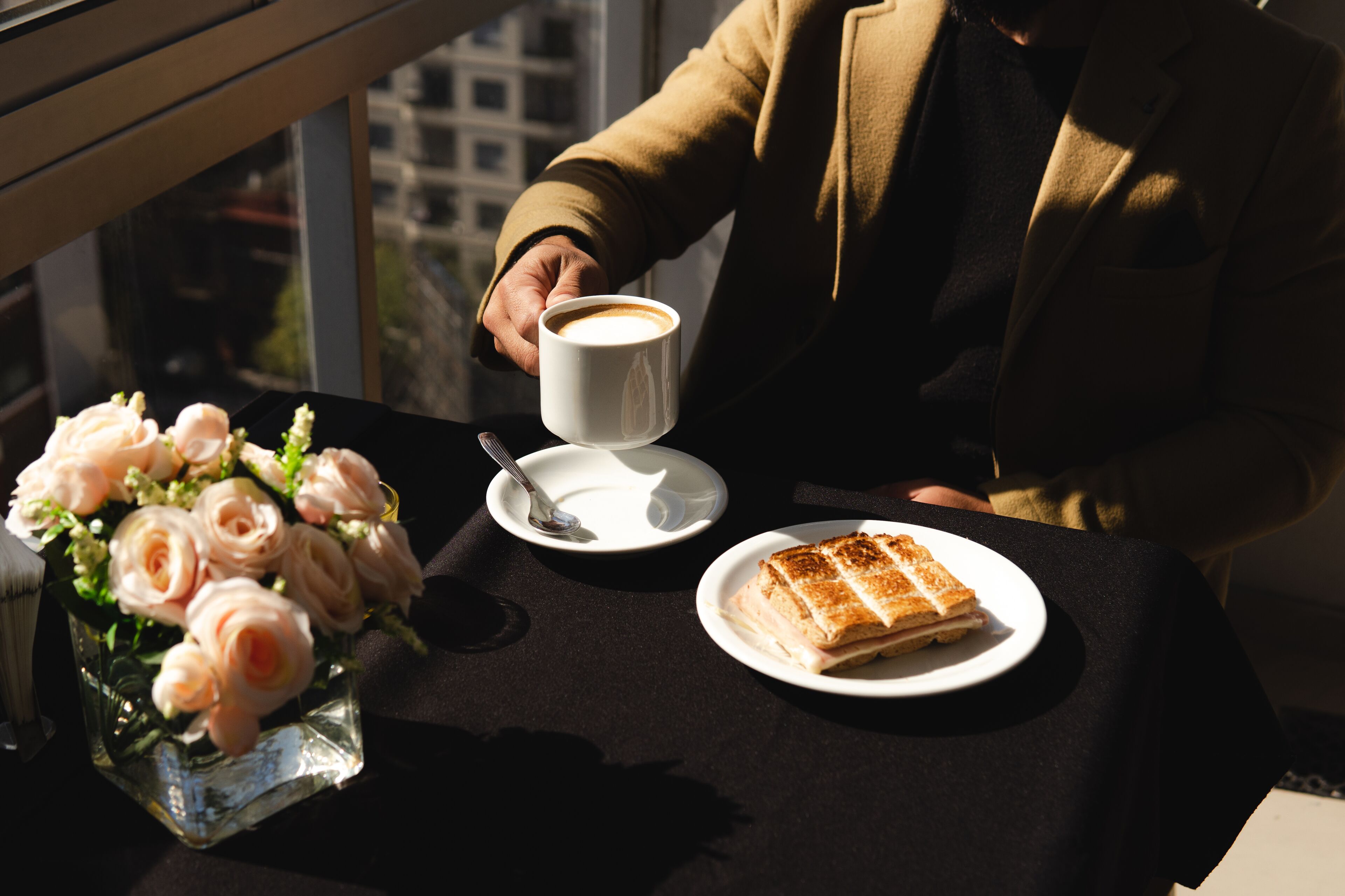 Une table avec une tasse à café blanche, une assiette contenant un sandwich et un vase rempli de roses roses.