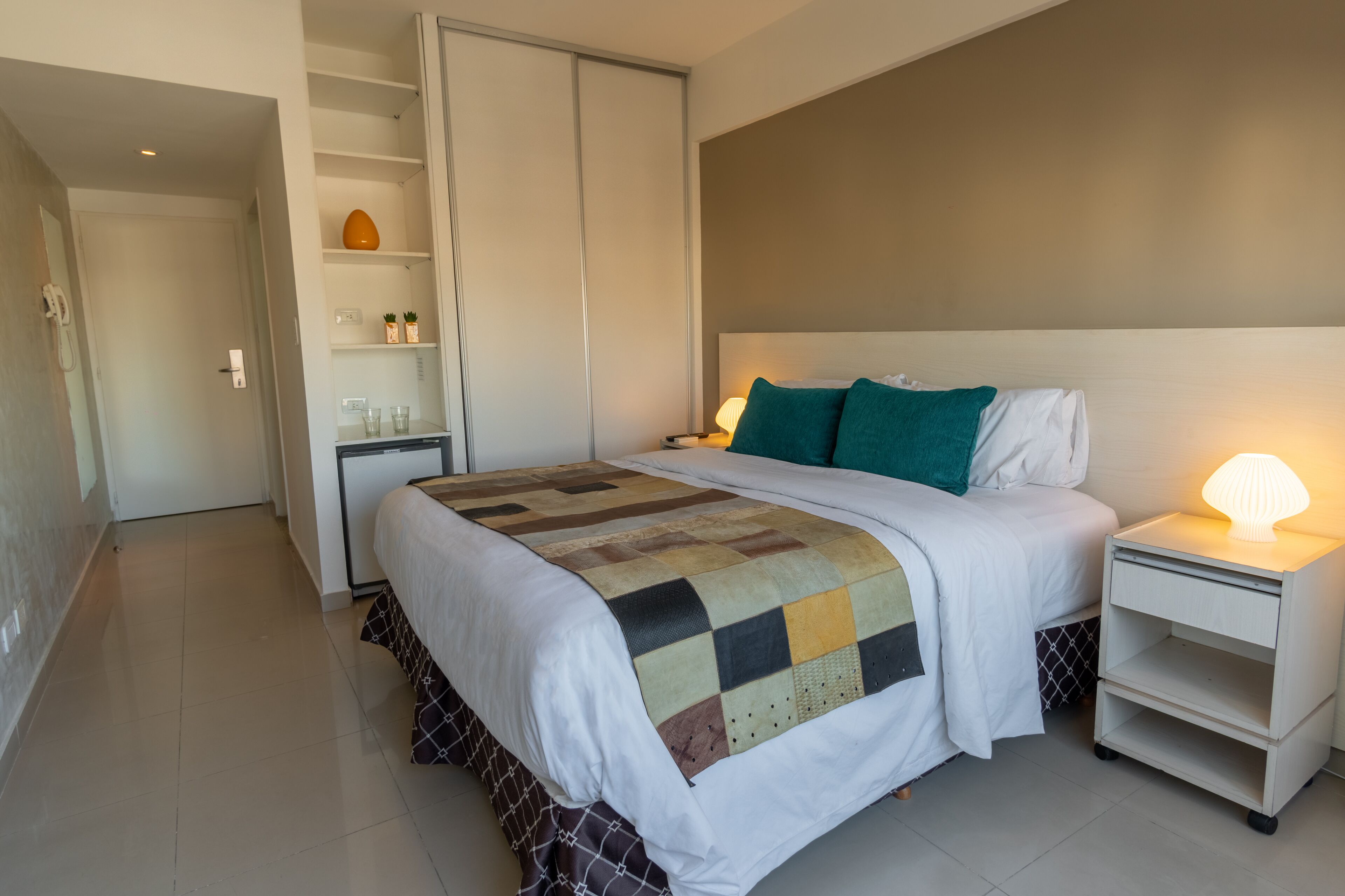 A neatly arranged bedroom with a bed, bedside tables, a wardrobe, and a shelf with decorative items.