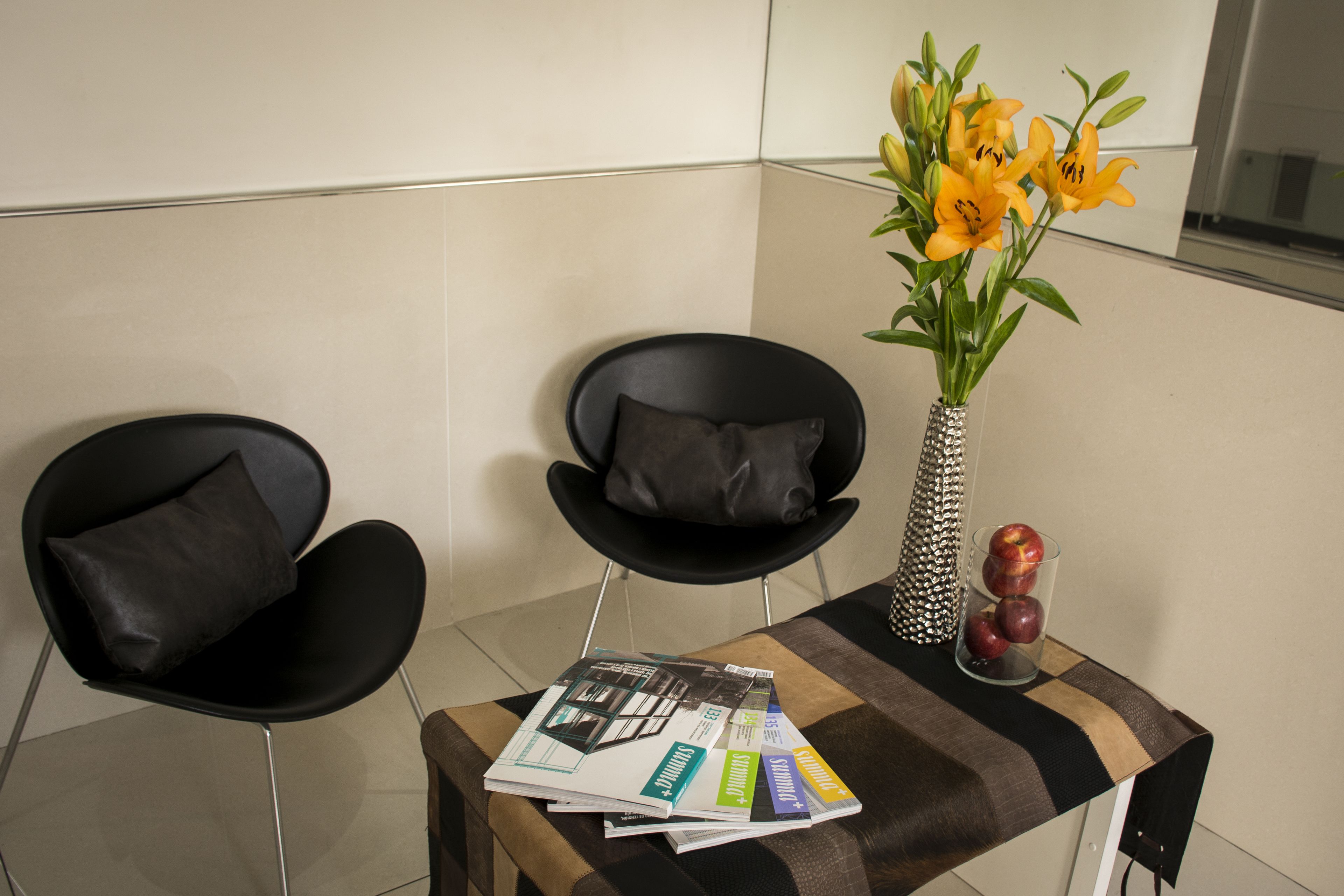 A modern room with two black chairs, a small table with magazines, a vase with yellow flowers, and a glass container with apples.