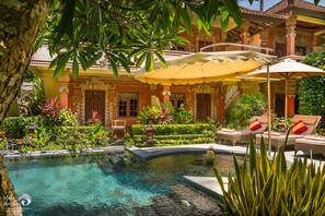 Uma área de piscina com espreguiçadeiras e guarda-sóis, rodeada por vegetação exuberante e um edifício tradicional com arquitetura ornamentada.