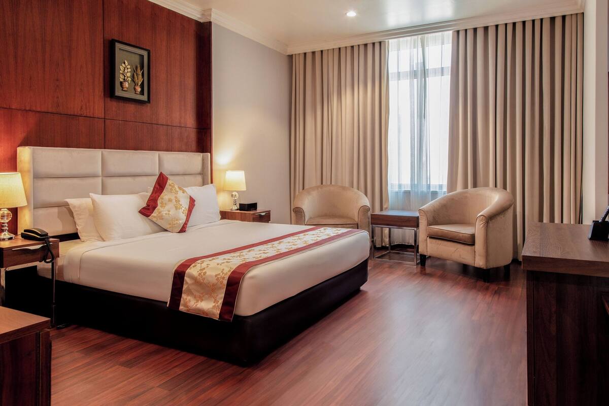 Une chambre d’hôtel avec un grand lit, deux tables de chevet avec des lampes, une chaise, une petite table et une grande fenêtre avec des rideaux.