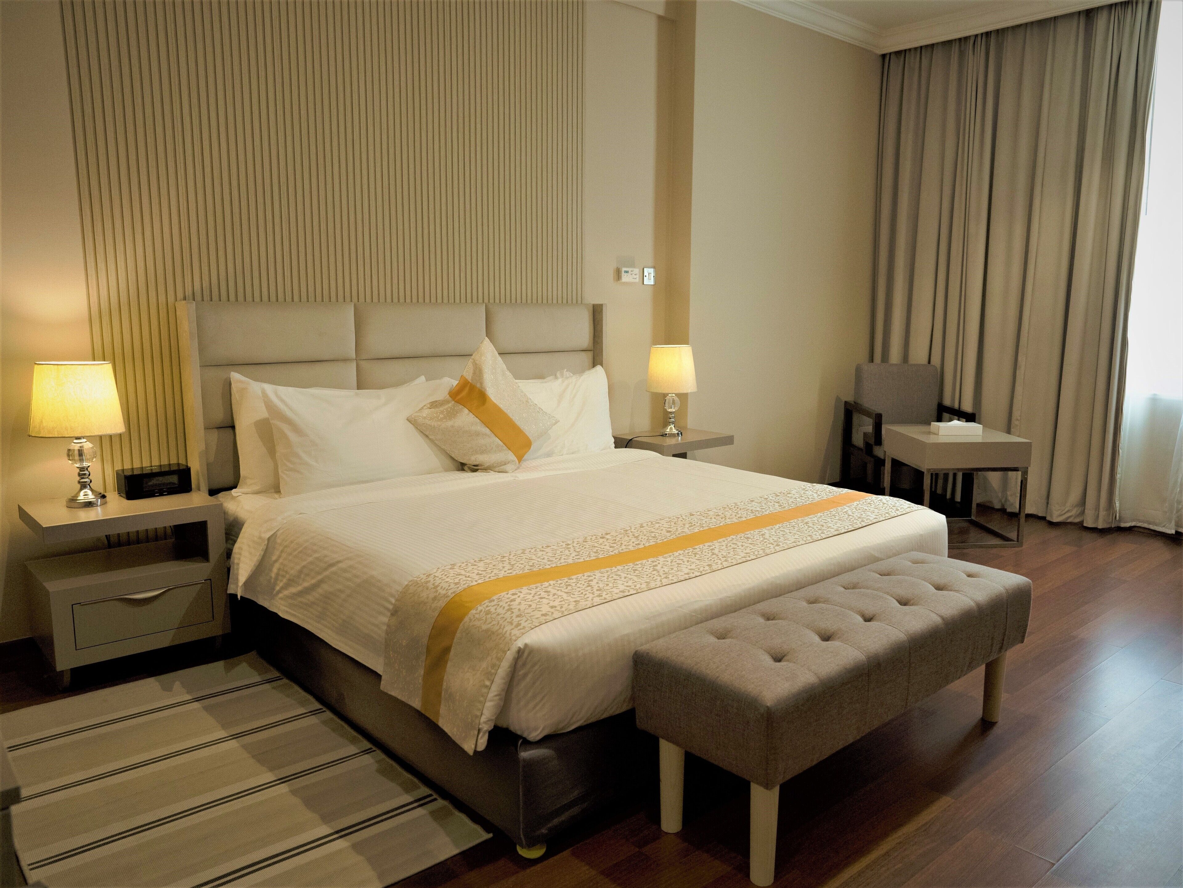 Una habitación de hotel con una cama grande, dos mesitas de noche con lámparas, una pequeña mesa, un banco y una ventana con cortinas.