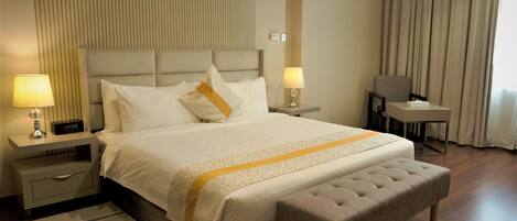 Una habitación de hotel con una cama grande, dos mesitas de noche con lámparas, una pequeña mesa, un banco y una ventana con cortinas.