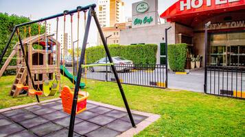 Un parque infantil con columpios y un tobogán, un estacionamiento y un hotel llamado "City Cafe" con un cartel.