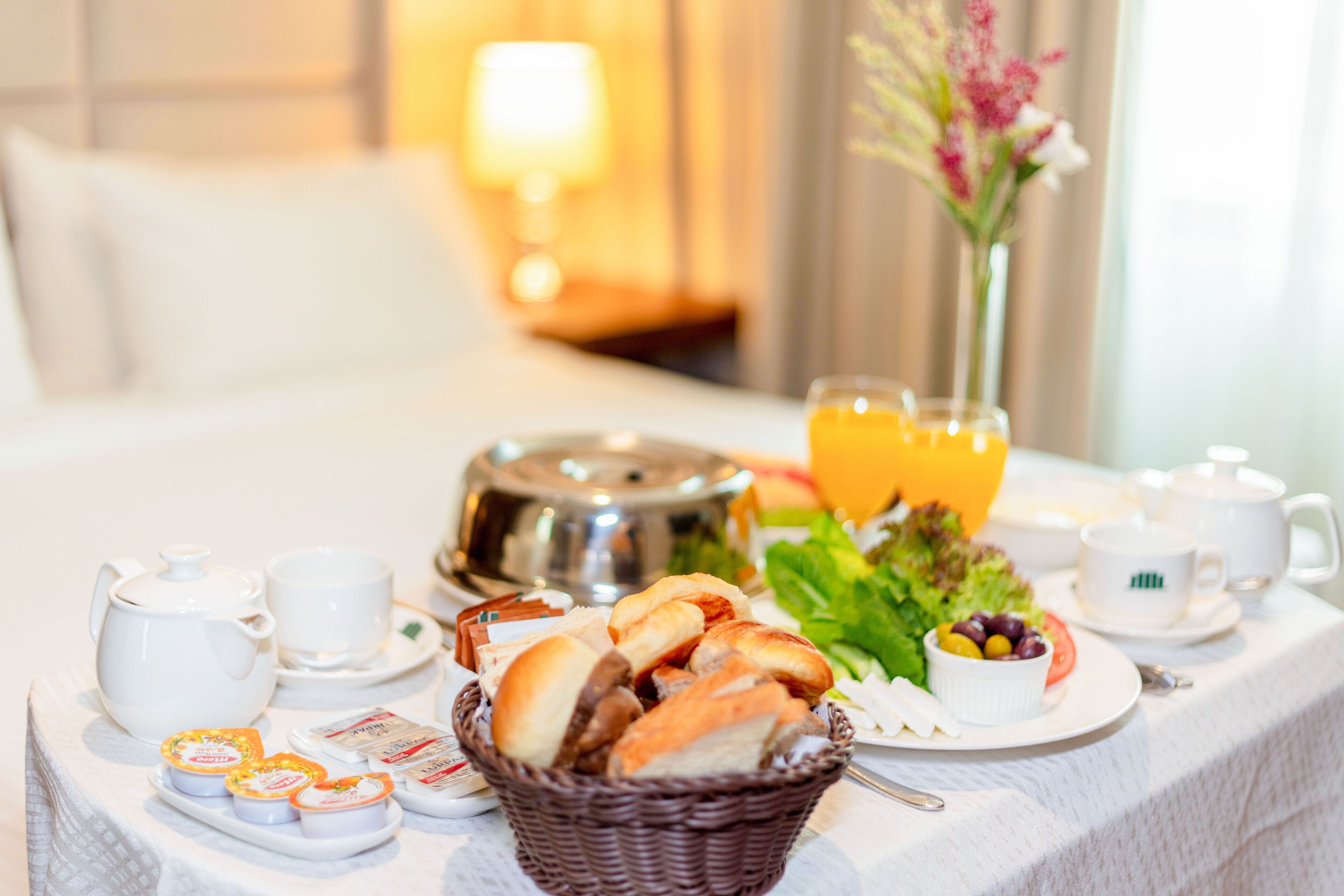 A breakfast setup on a bed with a basket of bread, a plate of olives, a glass of orange juice, and a teapot.