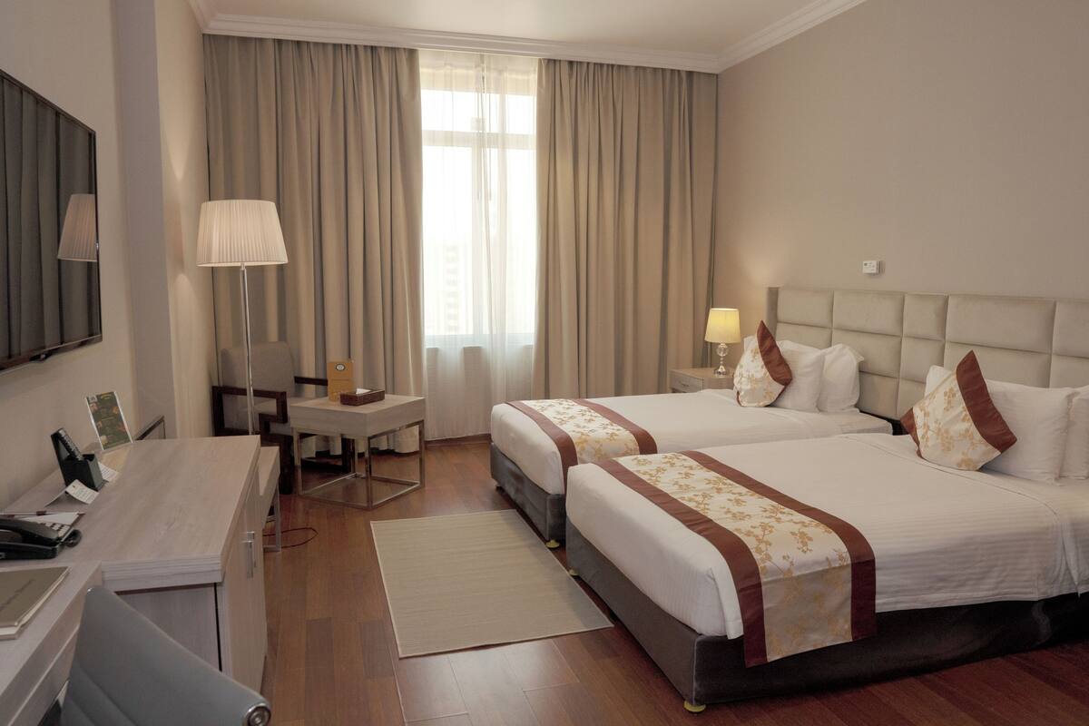 Une chambre d’hôtel avec deux lits, un bureau, une chaise, une lampe et une grande fenêtre avec des rideaux.