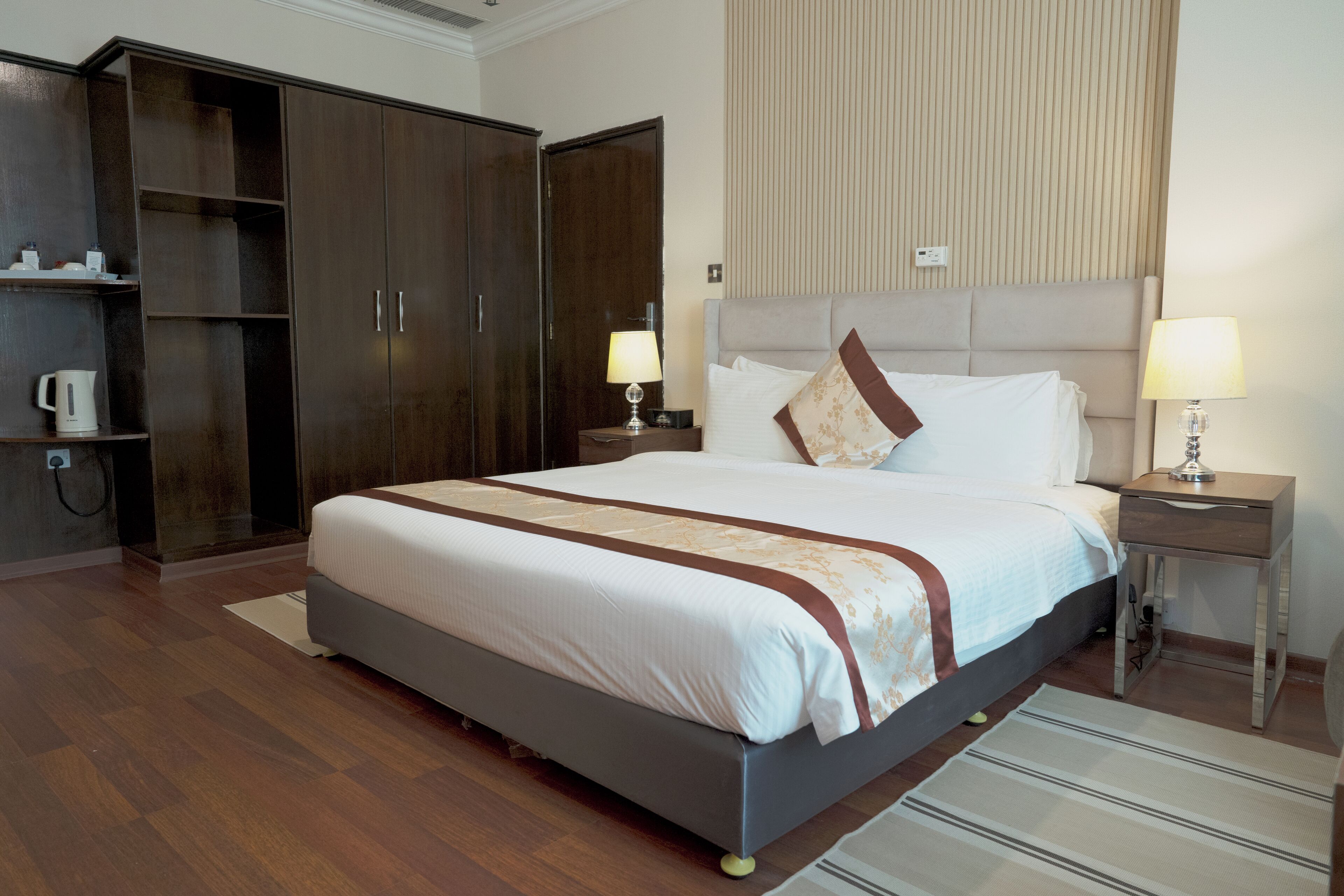 A hotel room with a large bed, a wardrobe, a bedside table with a lamp, and a small shelf with a kettle.