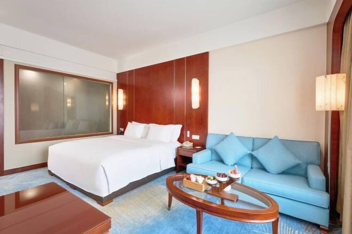 Une chambre d’hôtel avec un lit, un canapé bleu, une table basse en bois et une grande fenêtre.