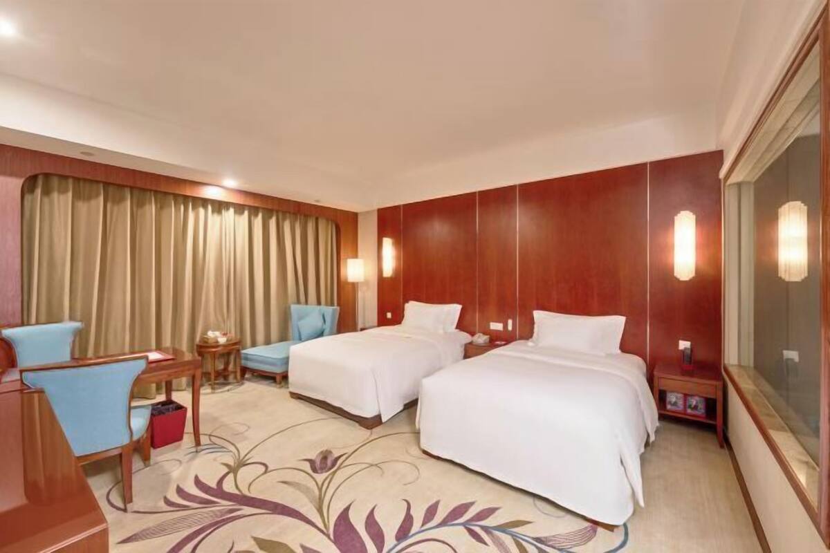 Une chambre d’hôtel avec deux lits, un bureau avec une chaise, une petite table et une grande fenêtre avec des rideaux.