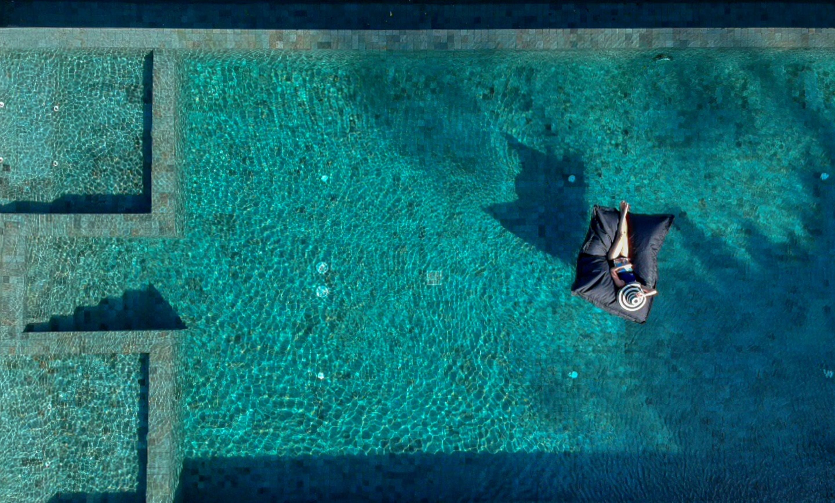 An aerial view of a swimming pool with clear water, a person lying on a lounge chair, and geometric tile patterns on the pool deck.