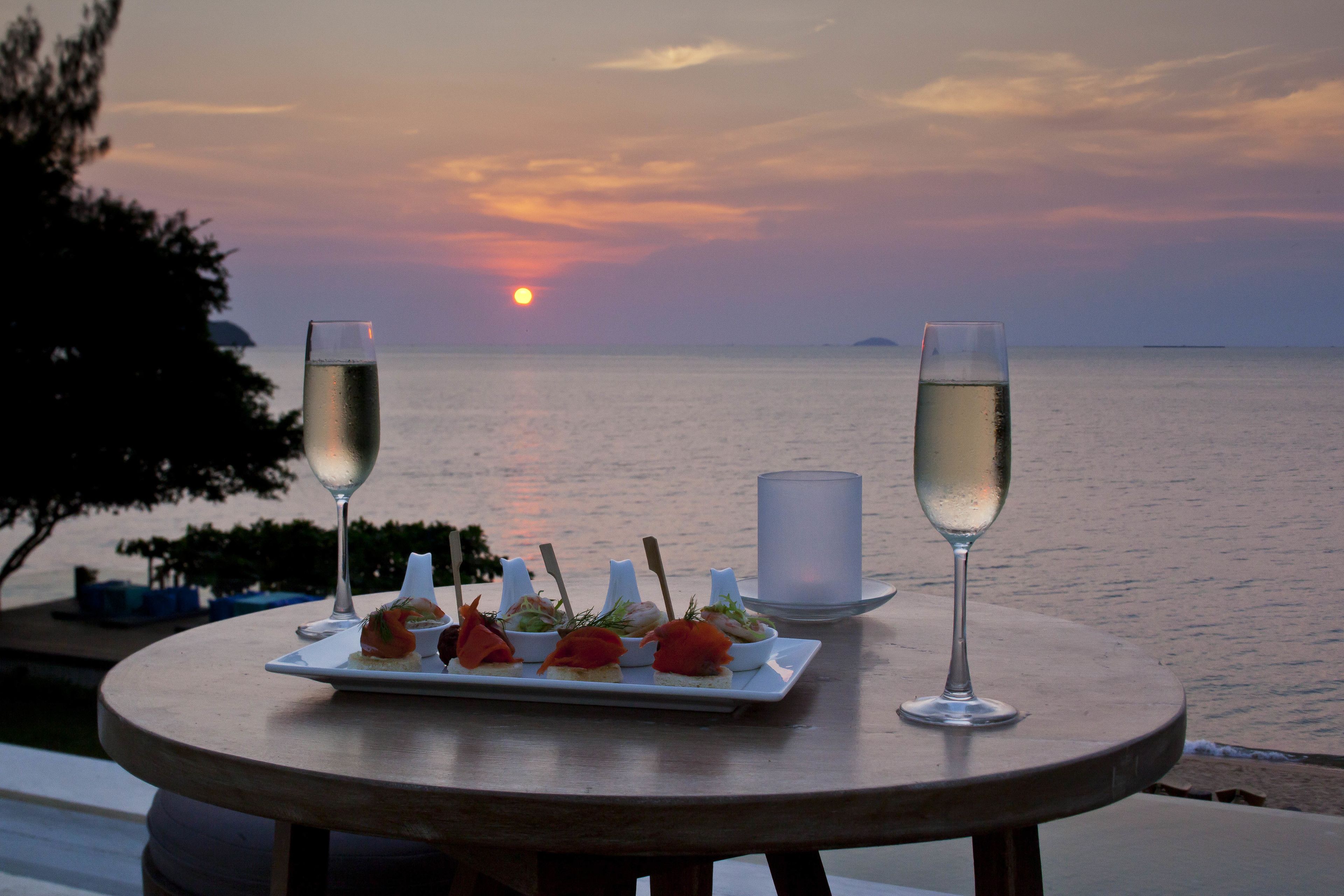 Un tavolo con un piatto di antipasti e due bicchieri di vino bianco, con vista sul mare al tramonto.