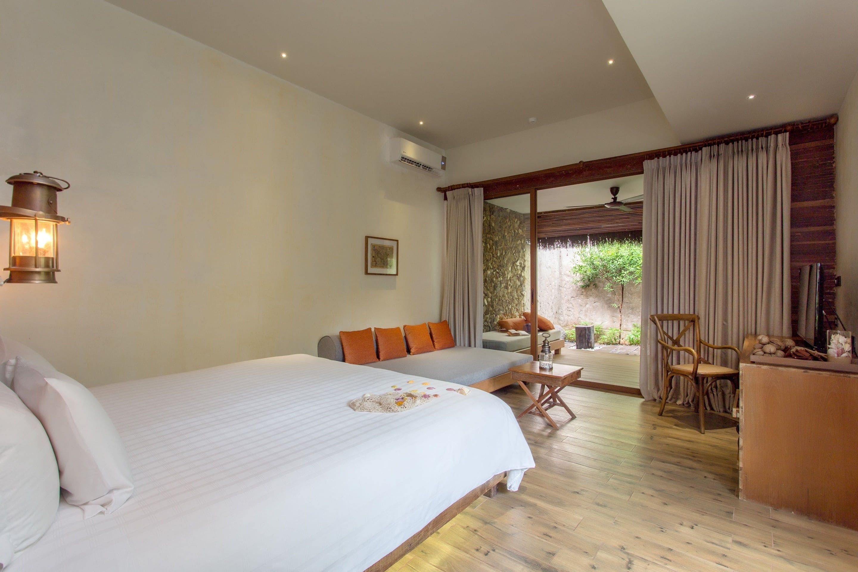Un dormitorio con una cama grande, un sofá, un escritorio y una silla. Cuenta con una ventana con cortinas, una lámpara de pared y un equipo de aire acondicionado.