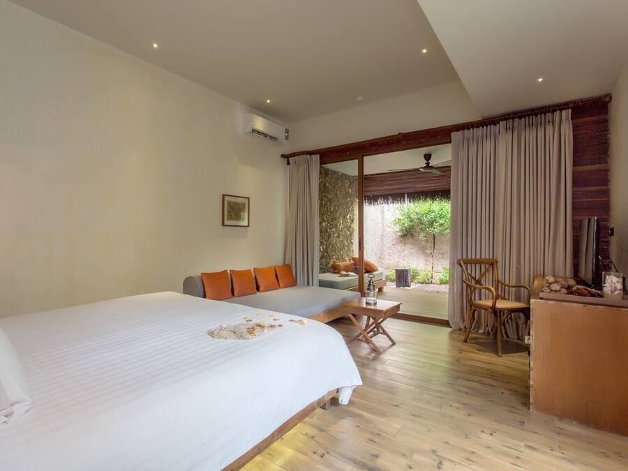 Un dormitorio con una cama grande, un sofá, un escritorio y una silla. Cuenta con una ventana con cortinas, una lámpara de pared y un equipo de aire acondicionado.