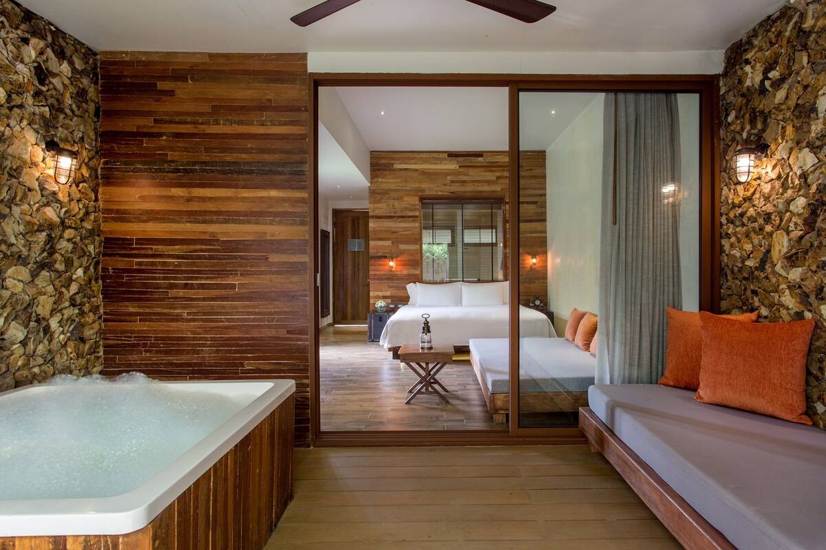 A modern hotel room with a stone wall, wooden paneling, a large bathtub, a sofa, and a bed with orange pillows.