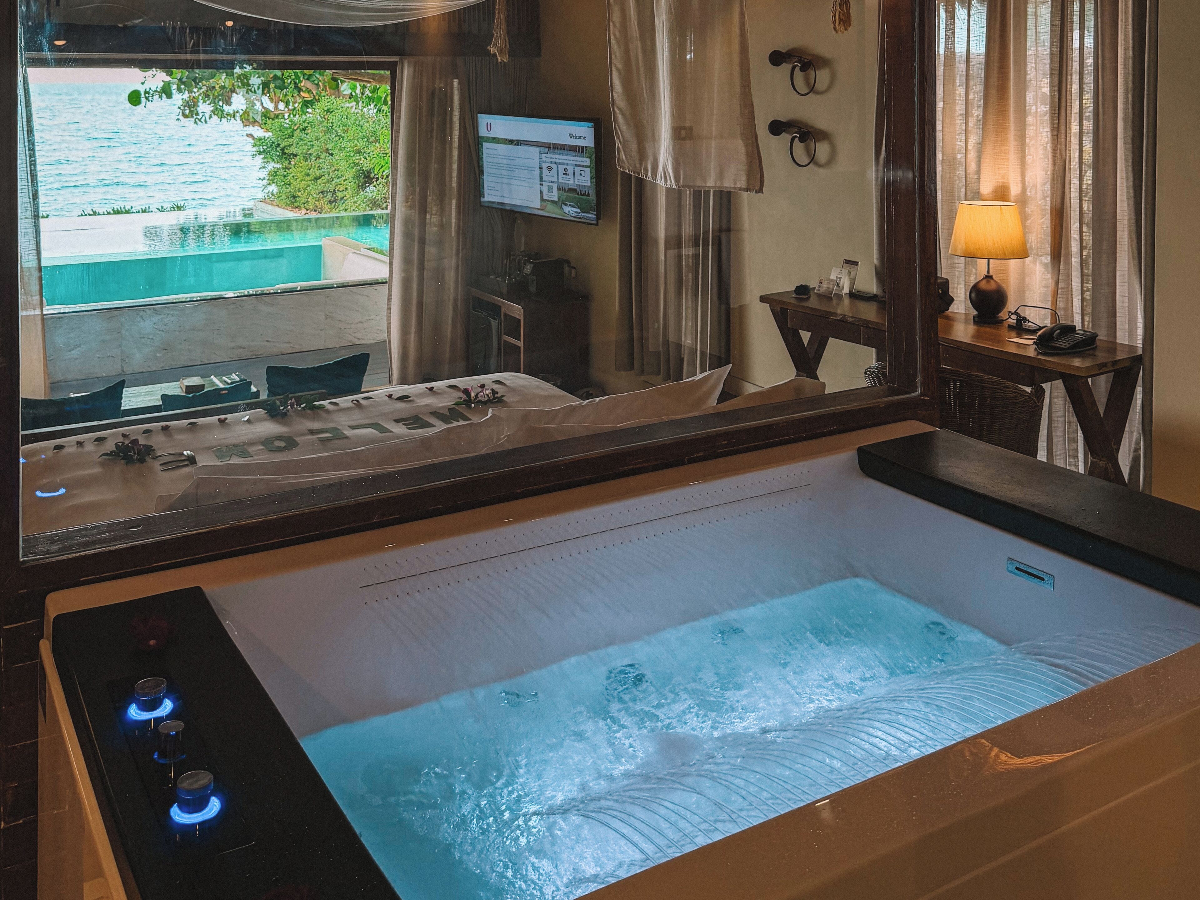Un baño con una bañera grande, un espejo, un lavabo y una ventana con vistas a una piscina y follaje.
