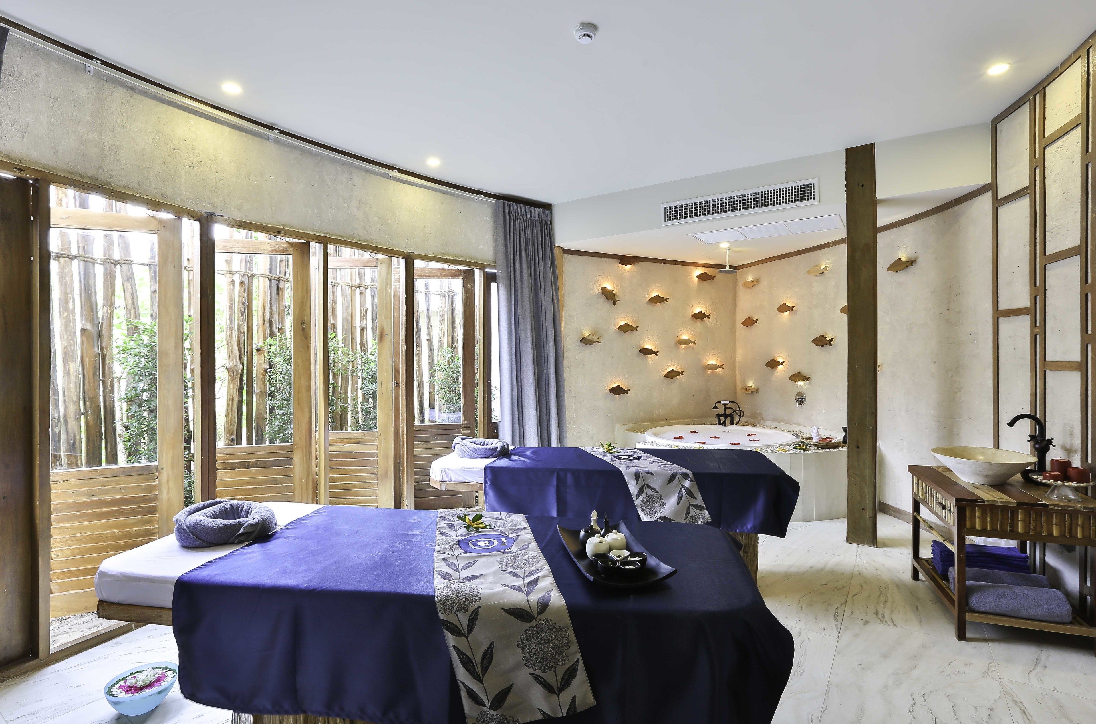 A spa room with two massage tables, a large window, a wall-mounted fish decoration, a sink, and a wooden shelf with towels.