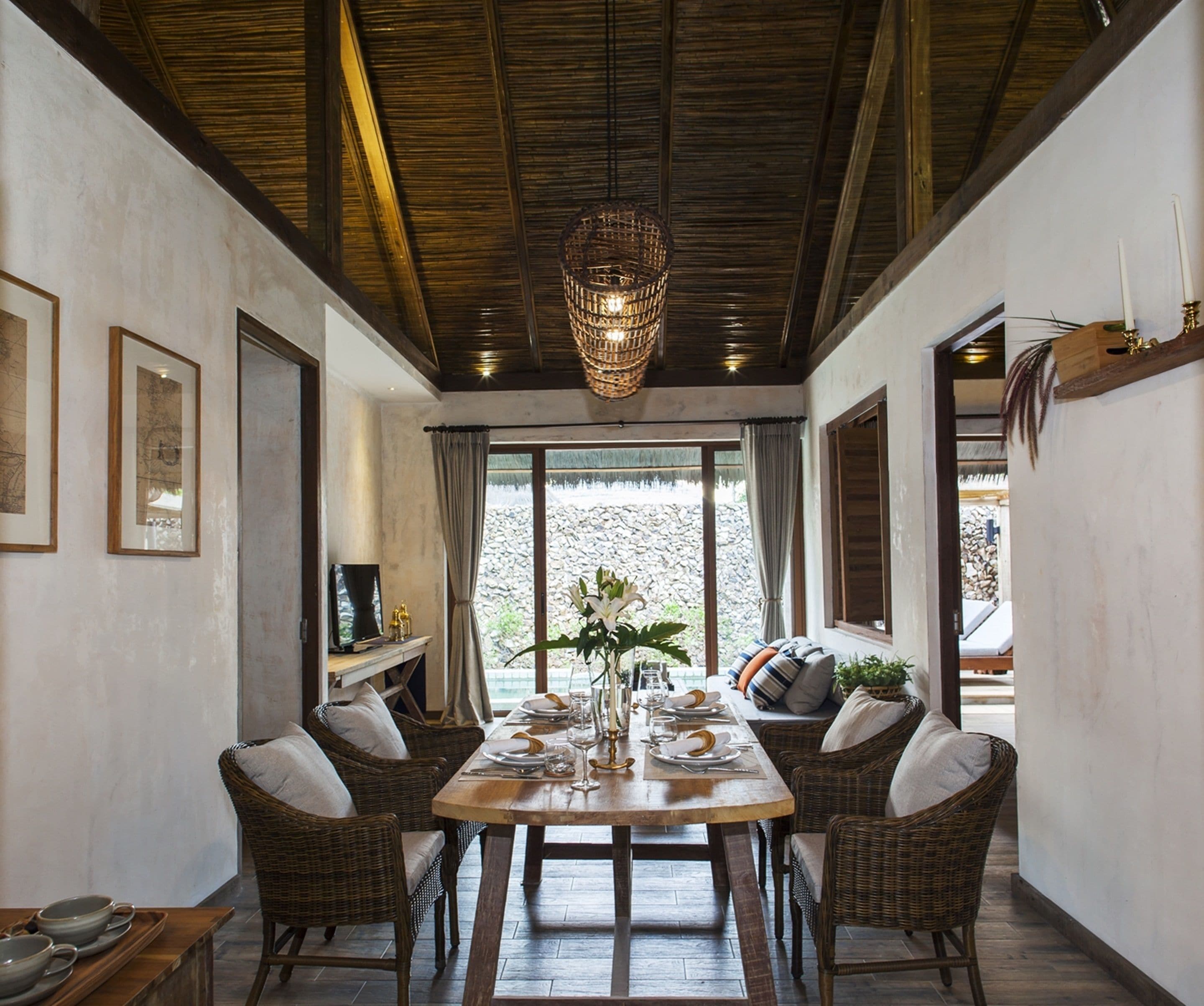 A dining area with a wooden table set for four, surrounded by wicker chairs. A large window provides a view of a rocky landscape.
