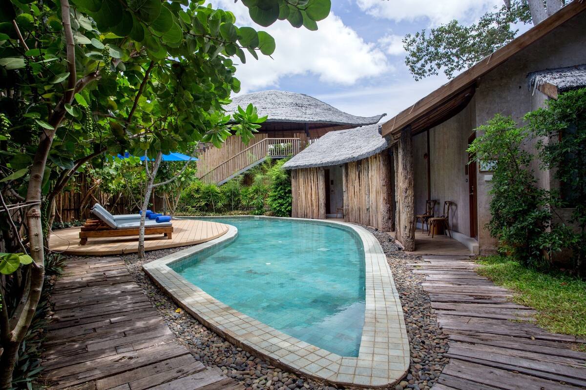 A pool area with a wooden deck, lounge chairs, and a thatched-roof structure.