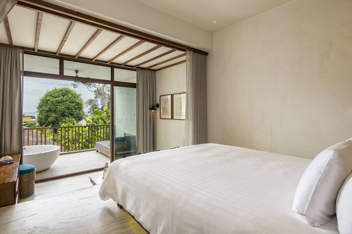 A bedroom with a large bed, a view of greenery through a sliding glass door, and a small seating area with a round table.