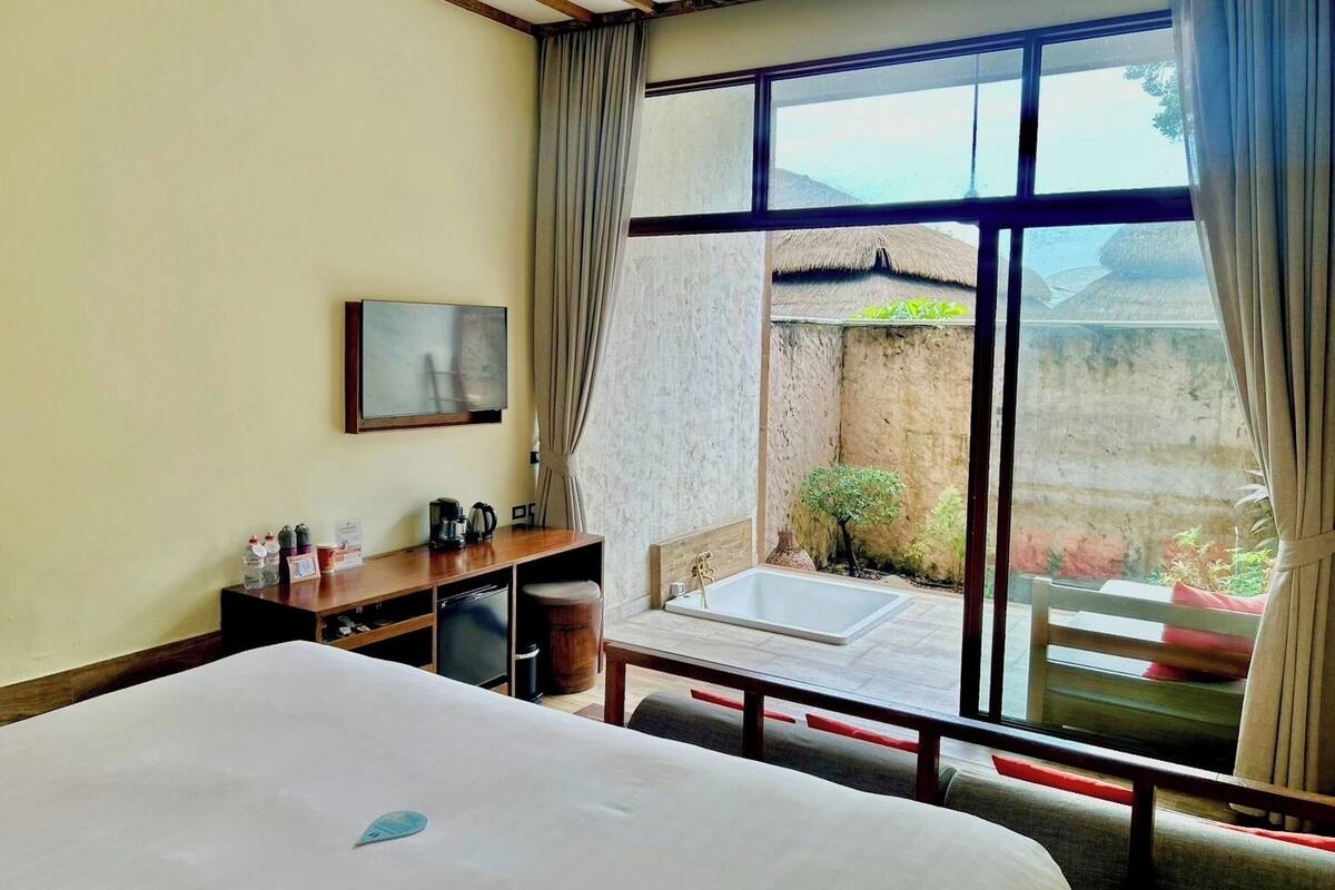 A hotel room with a bed, a desk, a chair, a small table, and a window with a view of a courtyard.