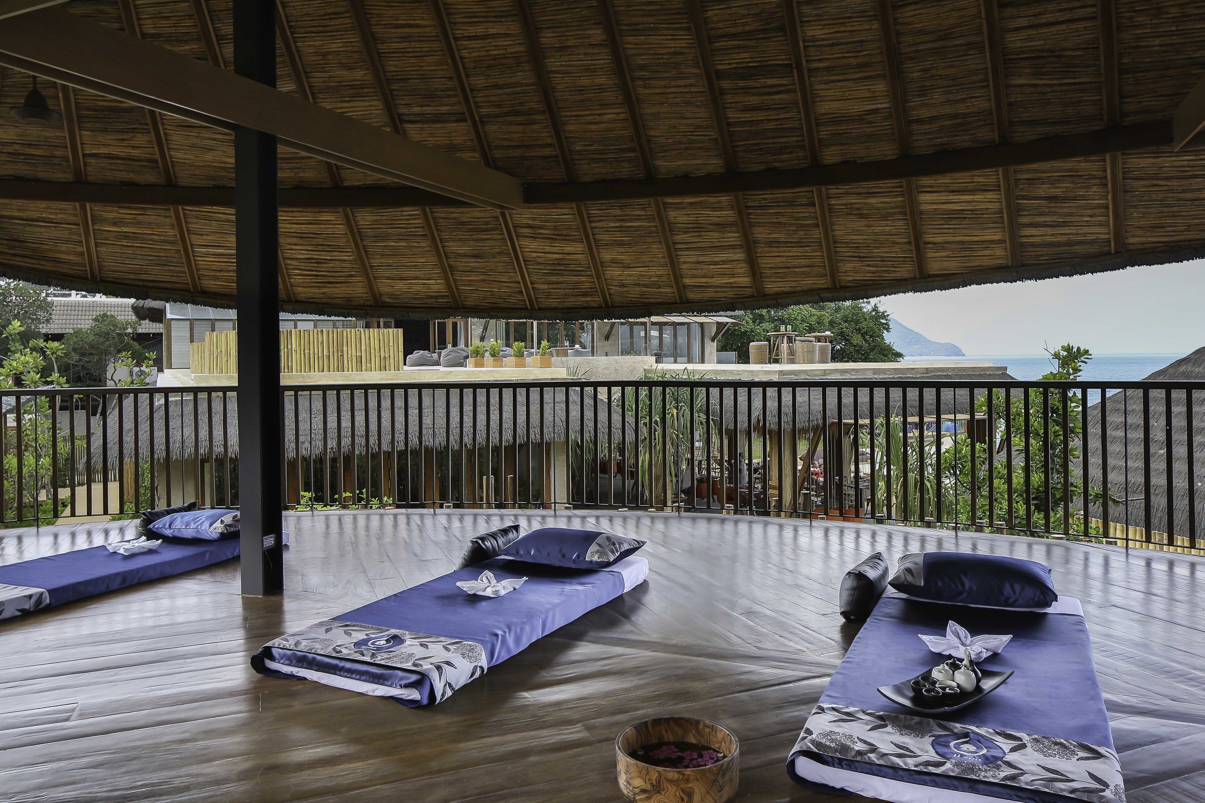 A serene outdoor lounge area with wooden flooring and thatched roof, featuring multiple reclining chairs with blue cushions and decorative pillows, set against a backdrop of lush greenery and a distant view of the sea.