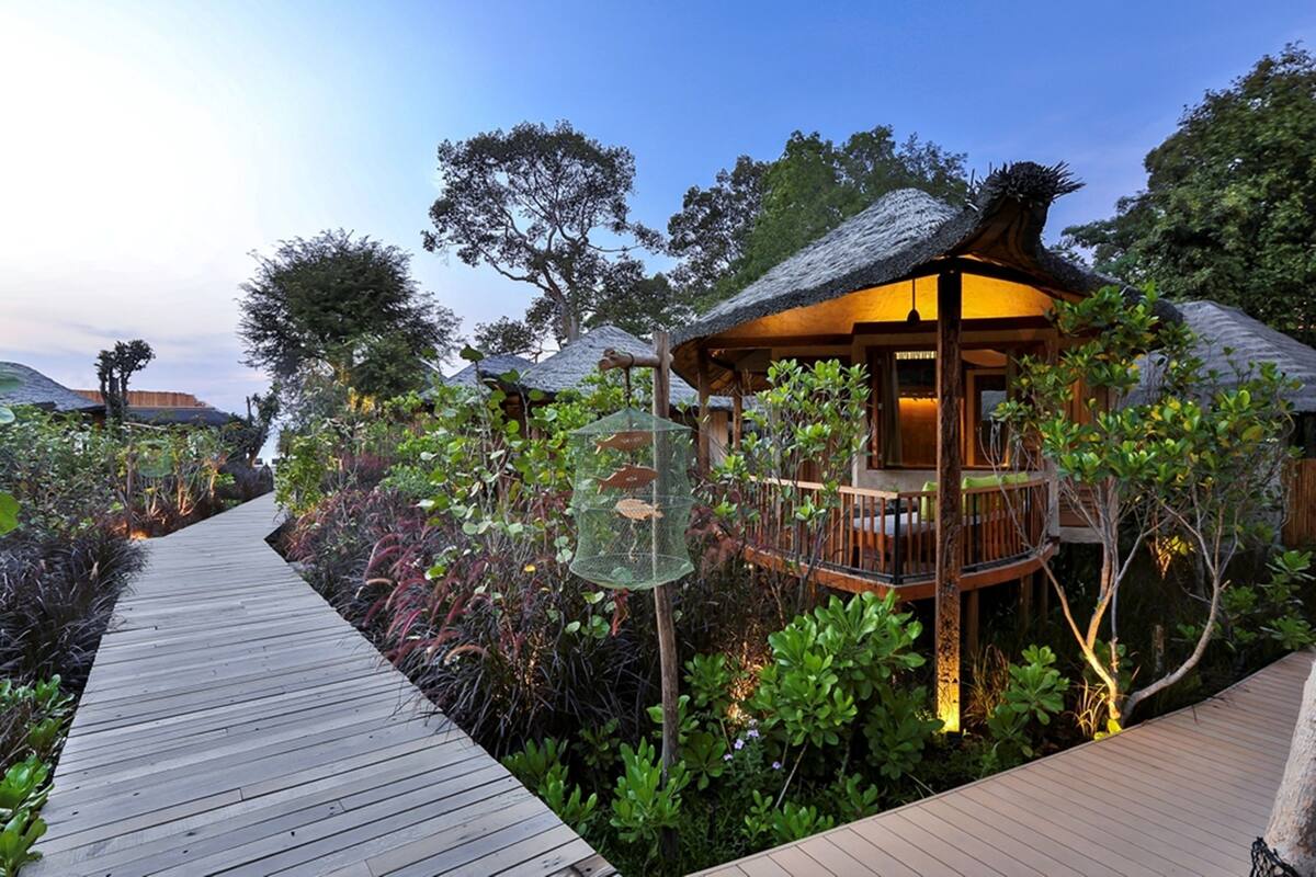 A wooden pathway leading to a thatched-roof structure with a balcony, surrounded by lush greenery.