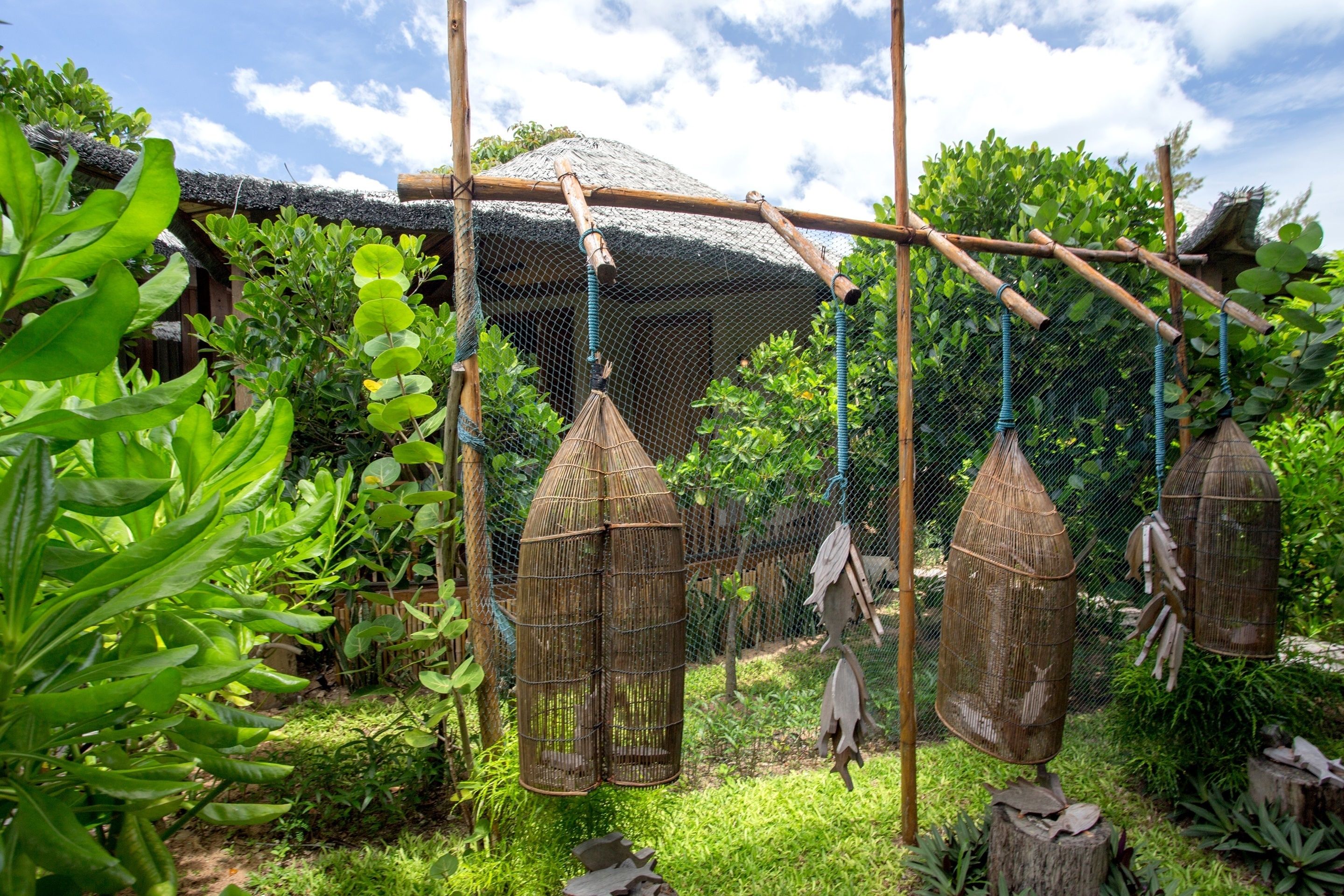 Un espacio al aire libre rústico con una estructura de bambú, jaulas de pájaros colgantes y abundante vegetación.