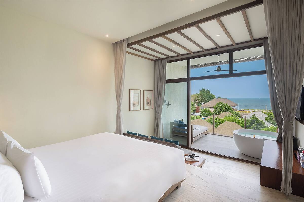 A bedroom with a large bed, a view of the beach, and a balcony with a bathtub.