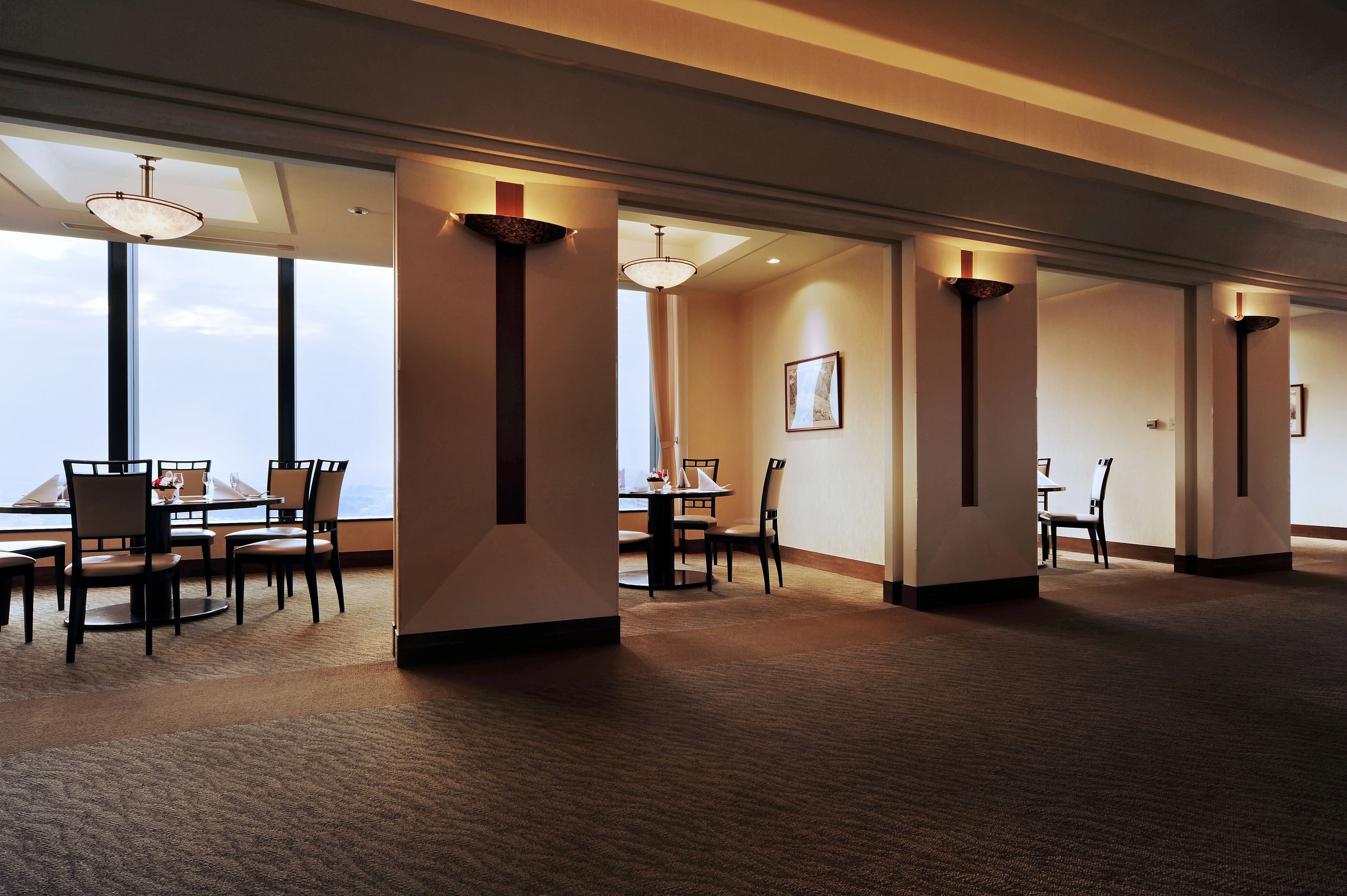 A hotel room with a large window, a dining area with chairs and a table, and a carpeted floor.