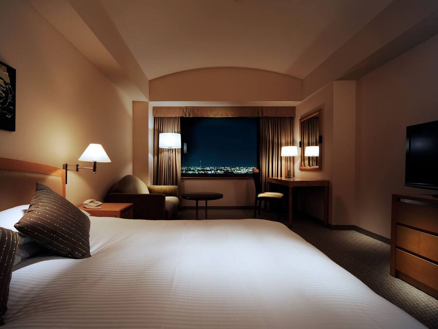 Una camera d'albergo con un letto grande, due lampade ai comodini, una scrivania con una sedia, una televisione e vista sulla città di notte.
