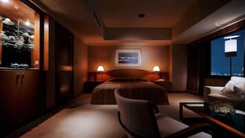 Ein Hotelzimmer mit einem Bett, zwei Nachttischlampen, einem Sessel, einem Schreibtisch, einem Fenster mit Blick auf die Stadt und einem an der Wand befestigten Regal mit Gläsern.