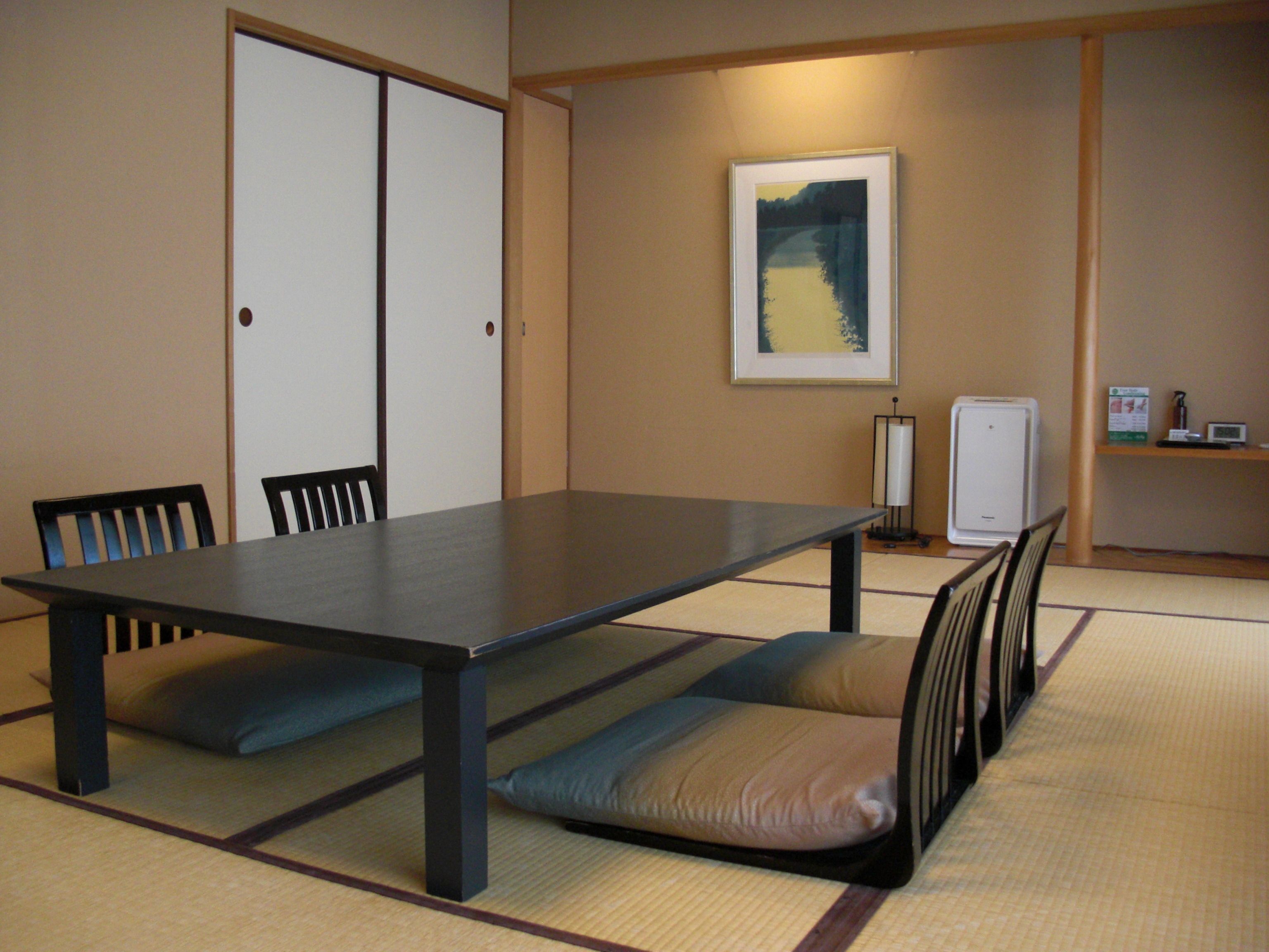 Un salón de conferencias con una larga mesa, sillas, esteras de tatami, un cuadro en la pared y un refrigerador pequeño.