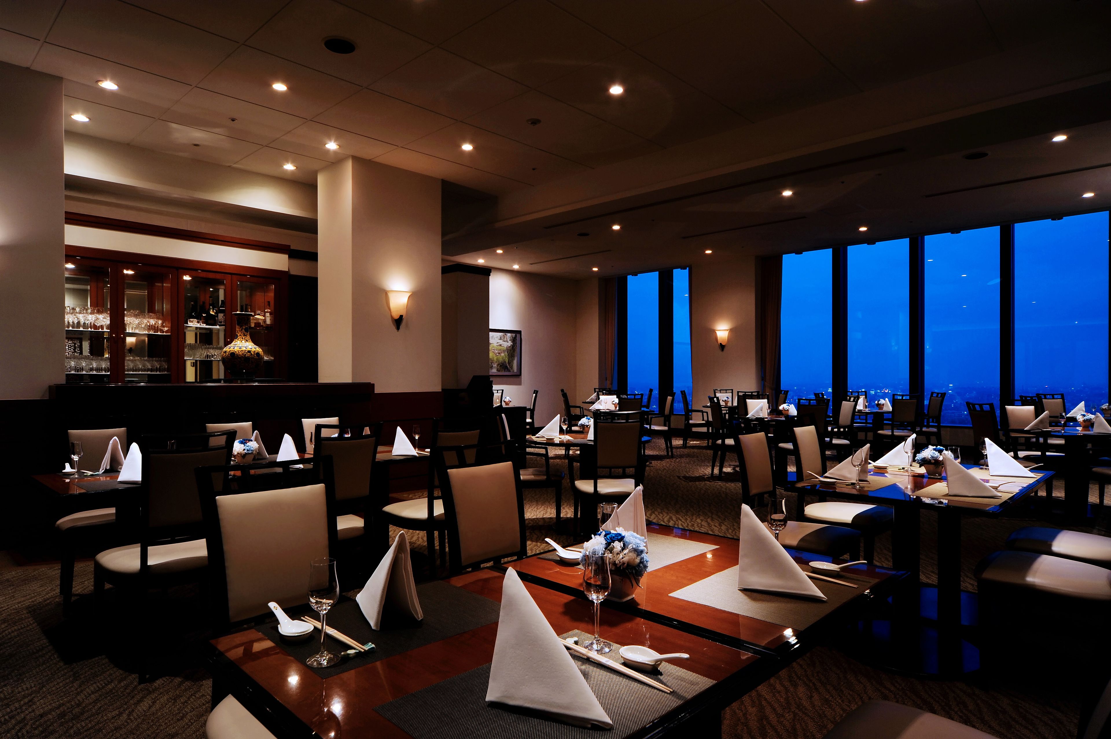 A restaurant with tables set for dining, a bar area, and large windows offering a view.