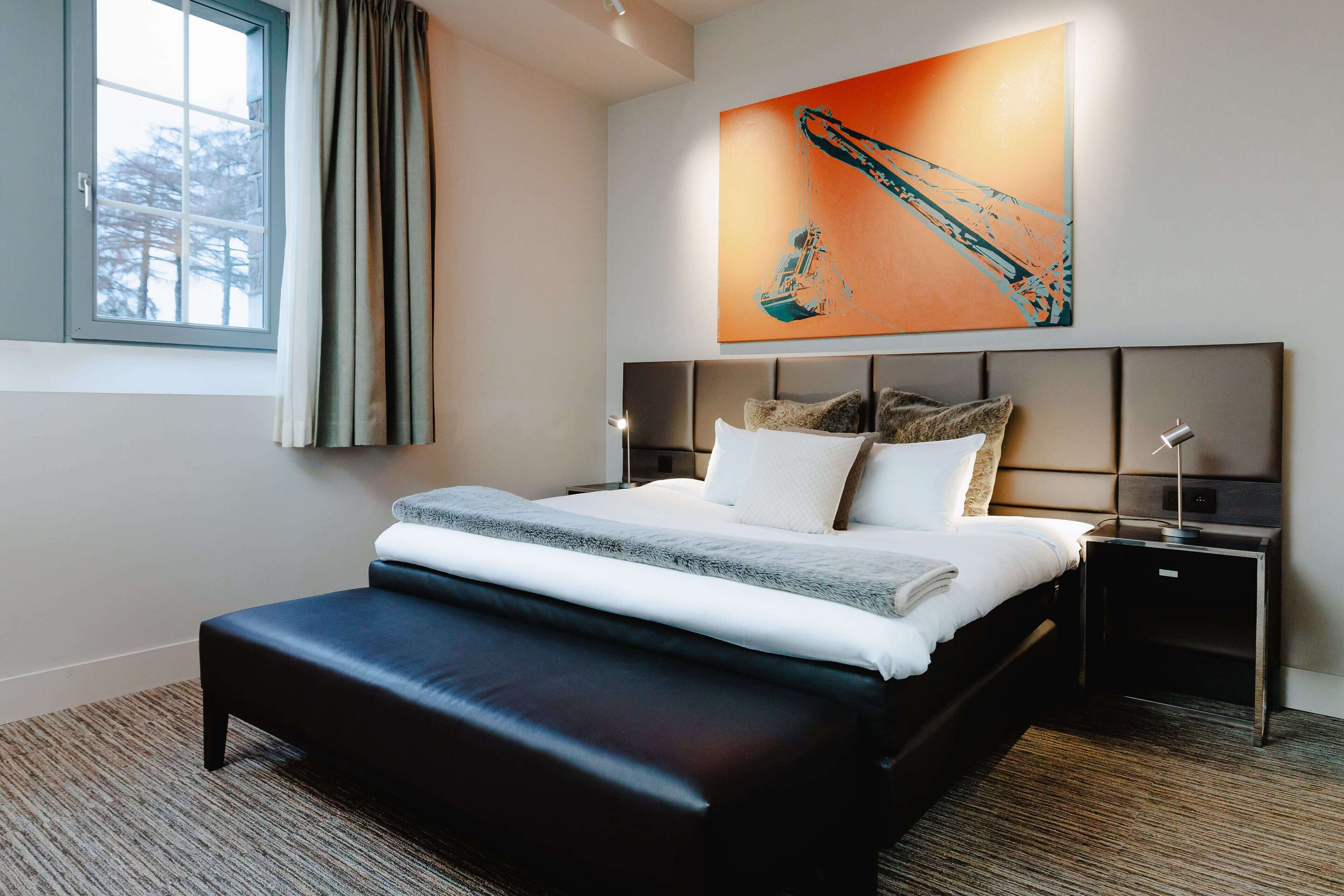 A modern hotel room with a large bed, a headboard with storage compartments, a bedside table with a lamp, and a window with curtains.