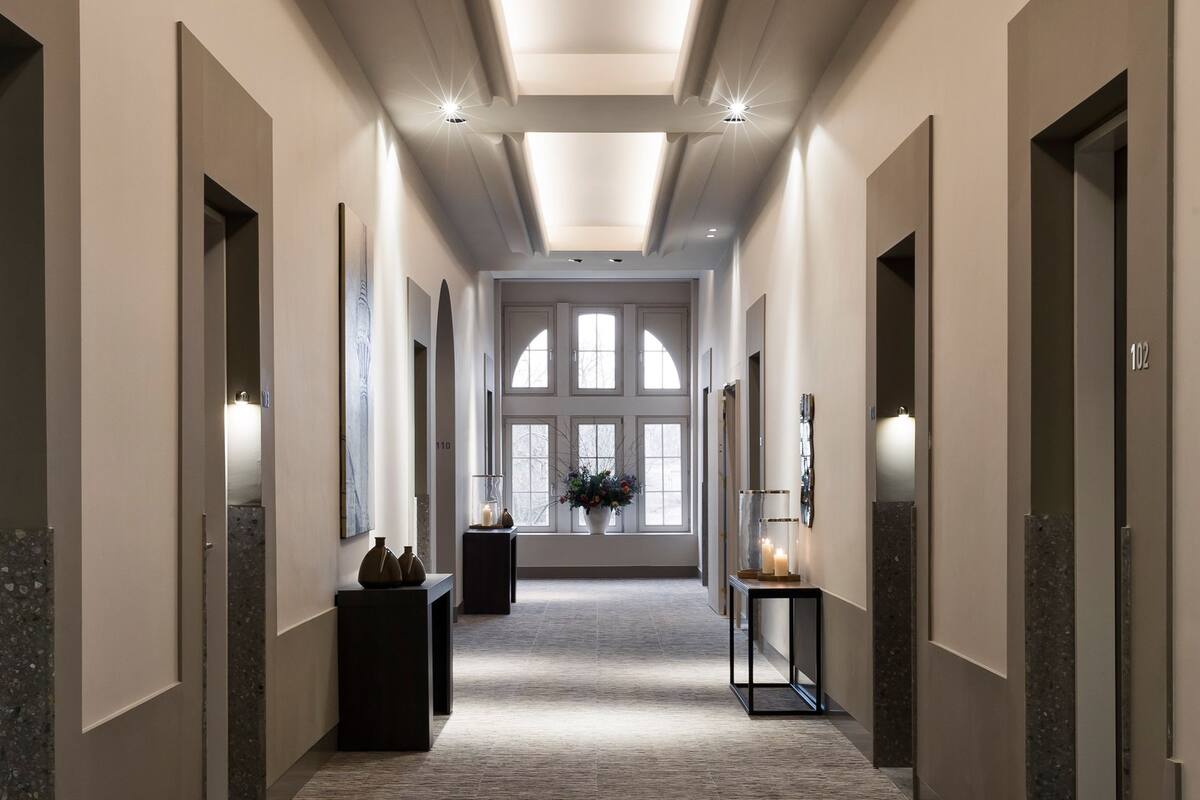 A long, narrow hallway with a carpeted floor, recessed lighting, and a large window at the end.