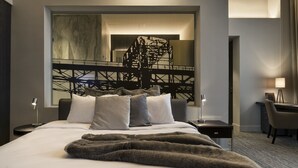 Habitación de hotel moderna con una cama grande, un escritorio y una silla. Se observa un espejo grande en la pared, y una escalera se ve en el reflejo.