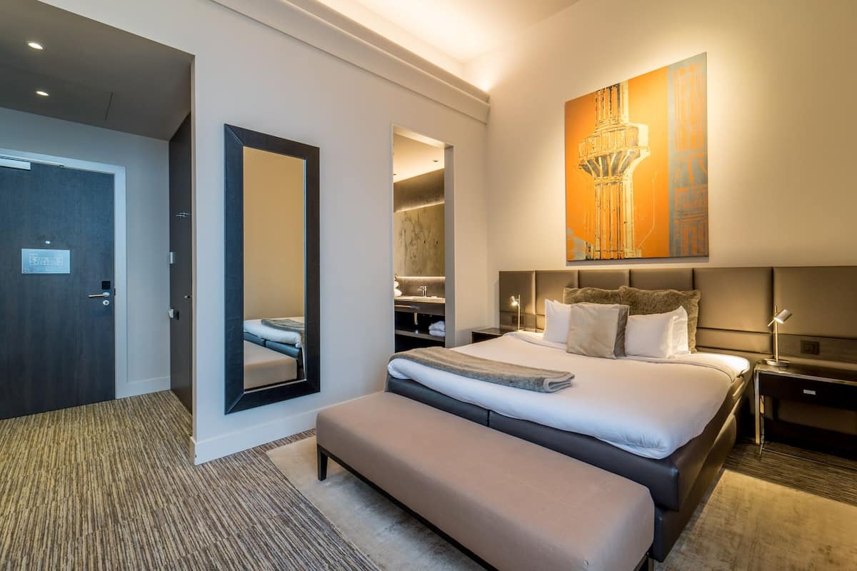 A modern hotel room with a large bed, a bench, a walk-in closet, and a large mirror.