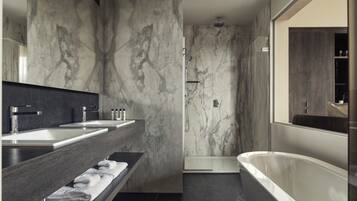 Une salle de bain moderne avec des murs en marbre, une douche à l’italienne, une baignoire sur pieds et un long meuble-lavabo avec un miroir.