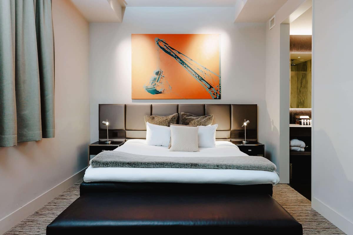 A modern hotel room with a large bed, a headboard with a built-in TV, a bedside table with a lamp, and a wall-mounted artwork.