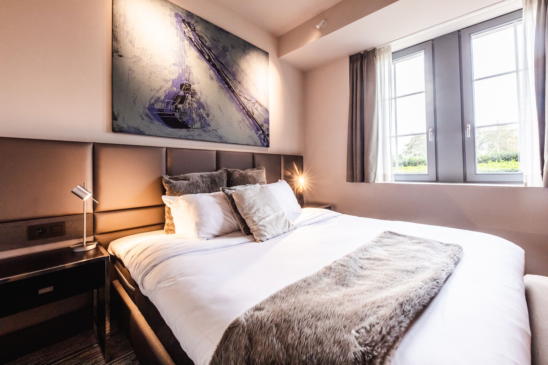 A modern hotel room with a large bed, a headboard with built-in lighting, a nightstand, and a painting above the bed.