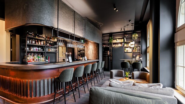 Un moderno área de bar con una barra curva, varios taburetes y estantes llenos de botellas y copas.