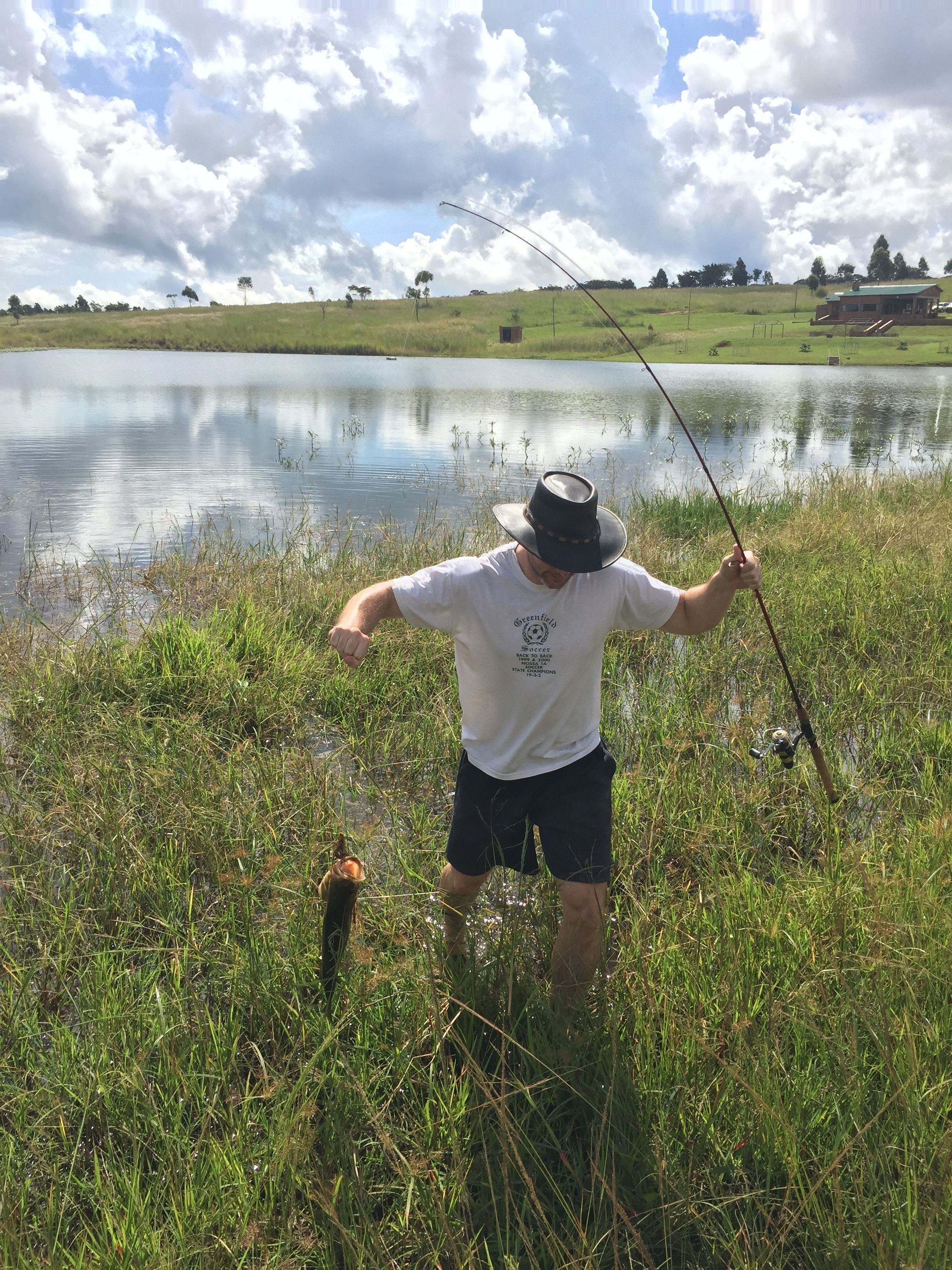 A person fishing by a calm lake with grassy banks and a clear sky.