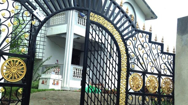 A gated entrance with ornate black and gold metalwork, a white building with a balcony, and a green object on the ground.