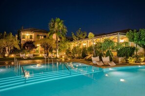 Una piscina con sillas de descanso y palmeras, rodeada de edificios con iluminación cálida.