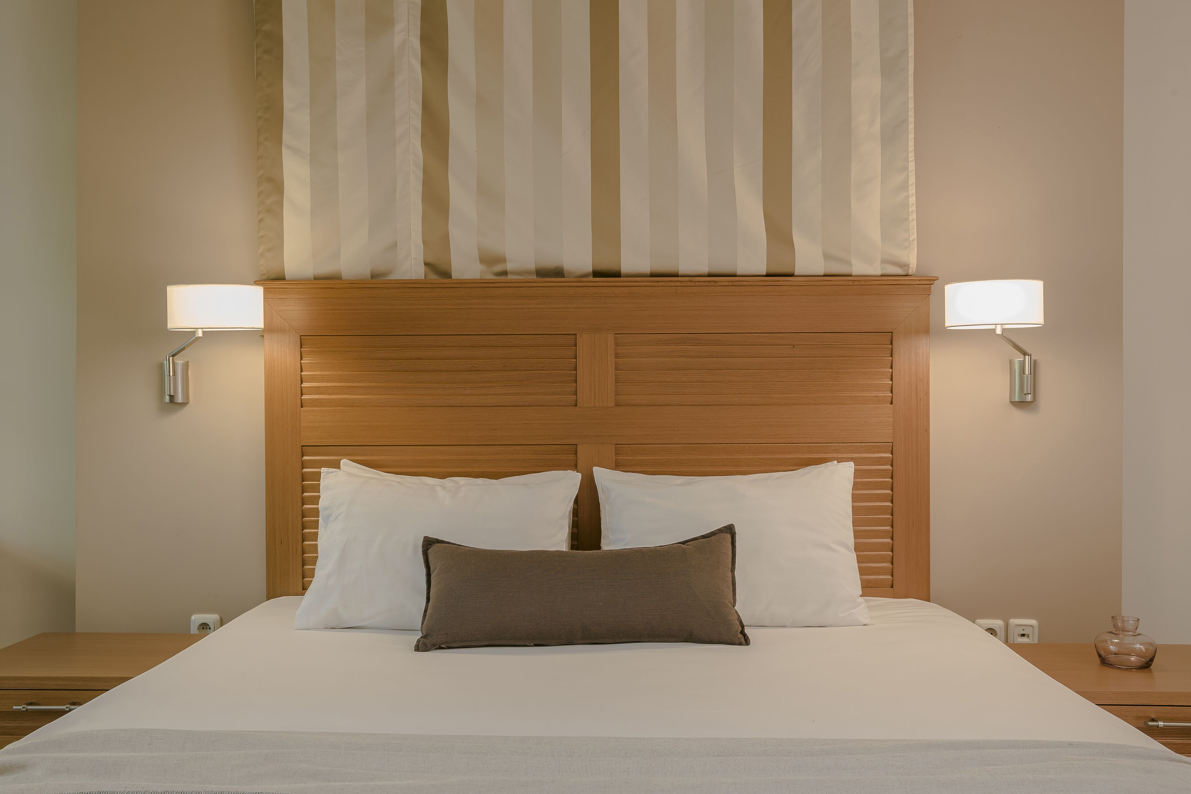 Una cama bien hecha con dos almohadas blancas y una marrón, flanqueada por dos lámparas de noche.
