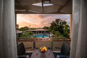 Un balcón con una mesa dispuesta para dos personas, un frutero y dos copas de vino. Al fondo, una piscina rodeada de vegetación y un edificio.