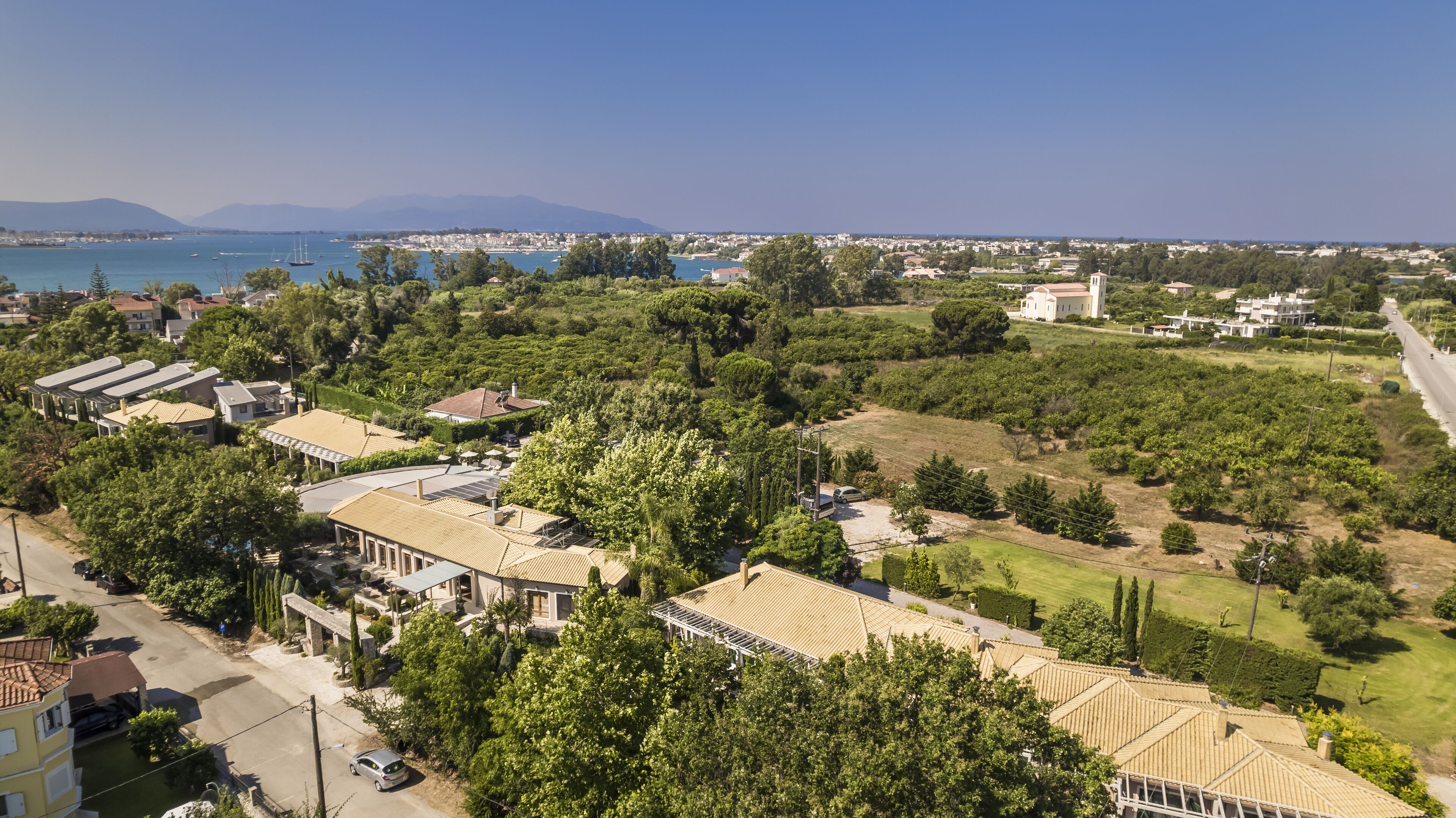 An aerial view of a coastal residential area with houses, greenery, and a clear view of the sea and mountains in the distance.
